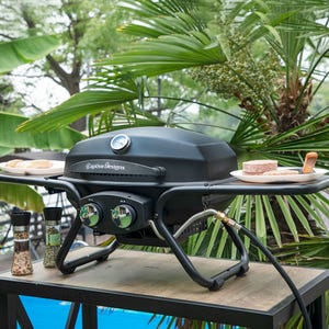 Portable gas grill on a table with a plate of raw meat next to spice grinders, set against a backdrop of tropical foliage.