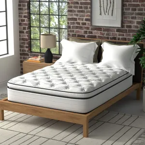 A white quilted mattress on a wooden bed frame, with two pillows, beside a nightstand with a lamp, in a room with brick walls.