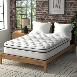A white quilted mattress on a wooden bed frame, with two pillows, beside a nightstand with a lamp, in a room with brick walls.
