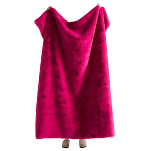 A person is holding up a large, bright pink, fluffy blanket. Only the shoes of the person holding the blanket are visible.