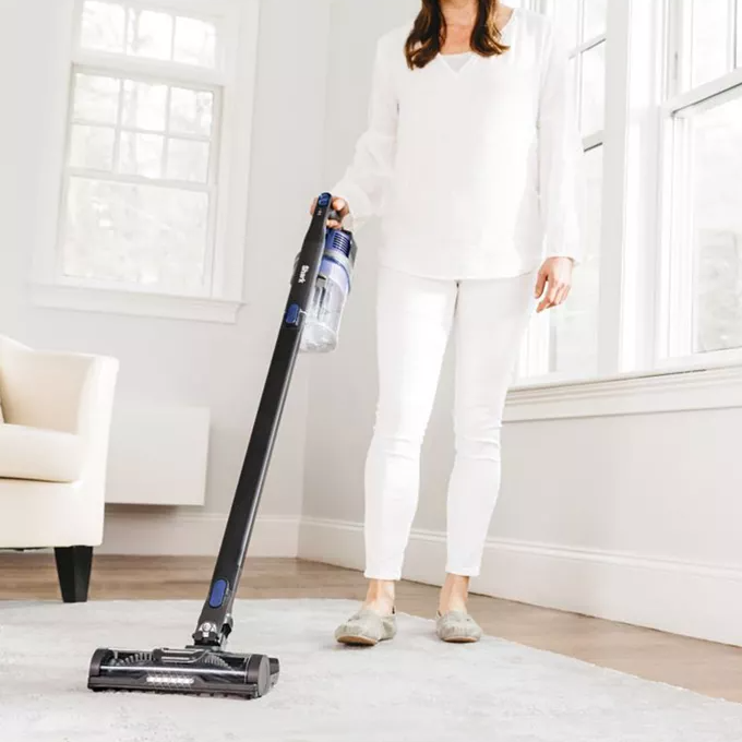 A person is holding an upright cordless vacuum cleaner while cleaning a carpeted room.
