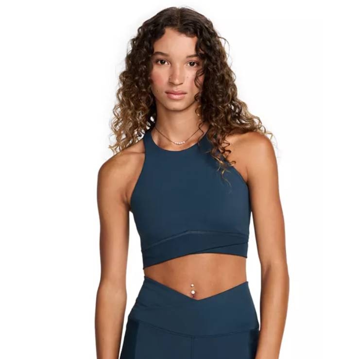 A woman is wearing a blue Nike high-neck sports bra with a fitted design, showcasing a sleek, supportive style for athletic activities. The top has a racerback and a cropped cut, providing a modern athletic look.