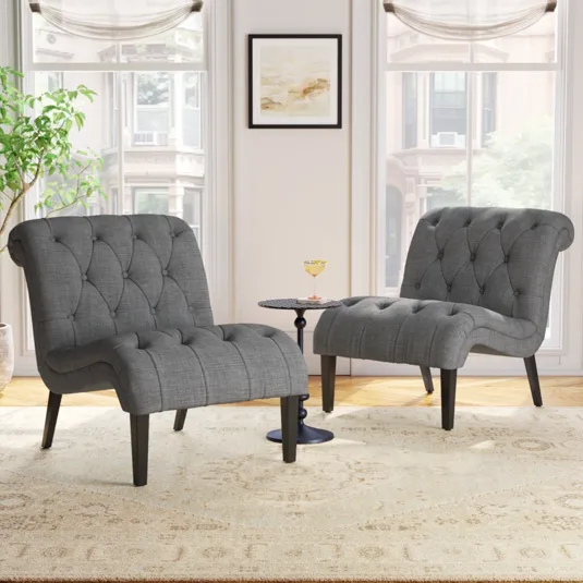 Two tufted gray lounge chairs with sloped arms are paired with a small round black side table, set on a light patterned rug in a bright, windowed room.