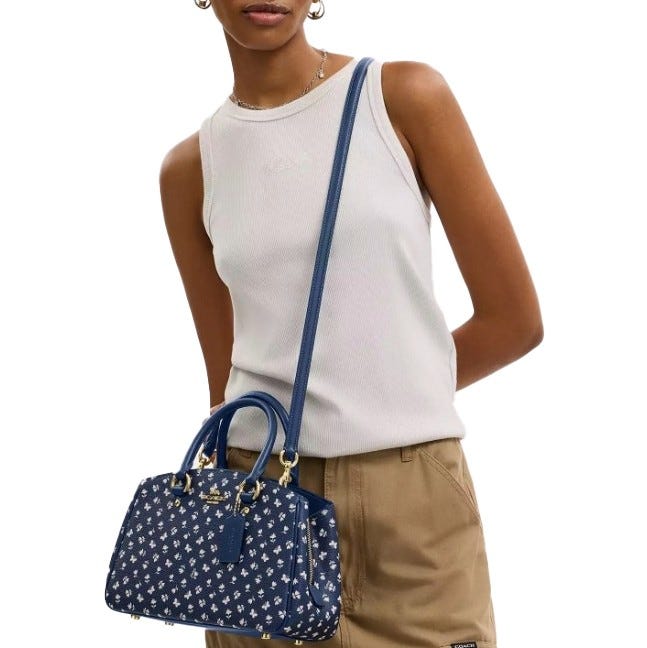 A person wears a sleeveless white top and beige pants, holding a blue floral patterned bag with a shoulder strap and top handles.