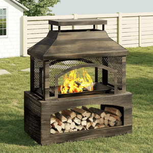 A metal outdoor fire pit with a mesh screen and log storage beneath, set in a backyard with grass and a white fence.