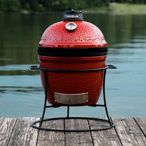 A small, red Kamado Joe Jr. charcoal grill with a ceramic body features a built-in temperature gauge on the lid, and it is supported by a sturdy metal stand on a wooden dock by the water.