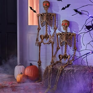 Two decorative skeletons with red light-up eyes are posed alongside pumpkins, with bats and cobwebs in the background for Halloween ambiance.