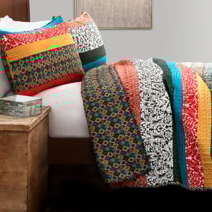 A colorful quilt and matching pillow shams feature a mix of geometric and floral patterns in various colors, including red, yellow, teal, and black, displayed on a bed with a rustic wooden nightstand nearby.