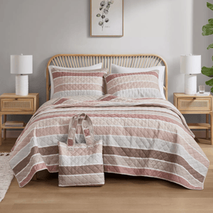 A cozy bedroom features a bed with a striped quilt and matching pillow shams, flanked by wooden nightstands and white lamps. A decorative wall art with botanical design hangs above, and a matching patterned tote bag rests on the bed.
