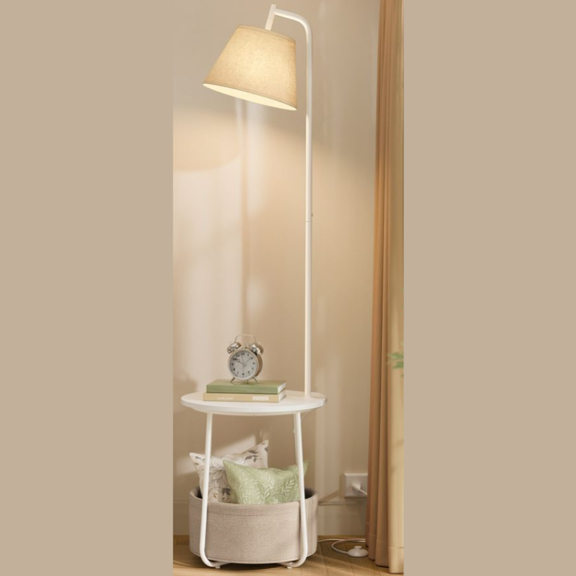 A tall floor lamp with a beige shade features an integrated side table holding a clock and books. Below, a basket contains decorative pillows.