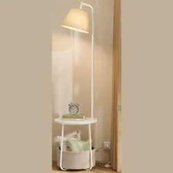 A tall floor lamp with a beige shade features an integrated side table holding a clock and books. Below, a basket contains decorative pillows.