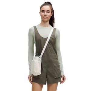 A person wears a sage green longsleeve top and olive romper, with a white crossbody bag holding a green water bottle.