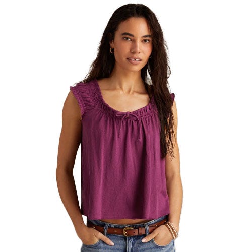 A woman is wearing a sleeveless, purple top with ruffled shoulders and a gathered neckline, paired with blue jeans and a brown belt.