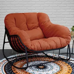 A cushioned rocking chair with an orange cushion and black frame is on a circular patterned rug in front of a white brick wall.