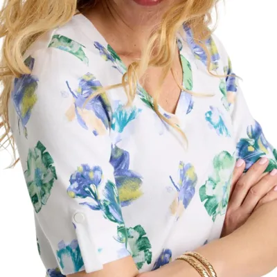 A white blouse with a floral pattern featuring shades of blue, green, and yellow, with rolled-up sleeves secured by a button detail. The person is also wearing gold bangles.
