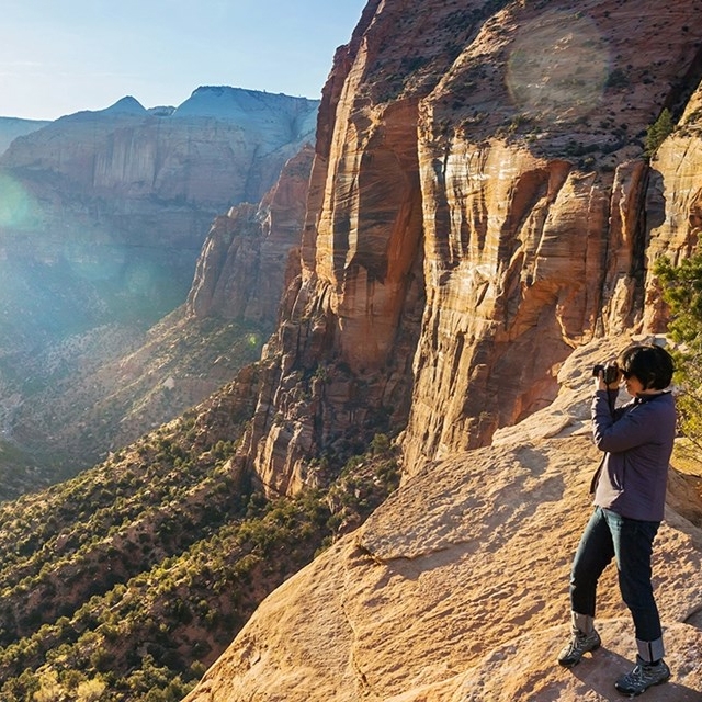 A person stands on a rocky cliff with a camera, capturing a view of a vast canyon landscape bathed in sunlight. The scene features rugged cliffs and a lush, sprawling valley.