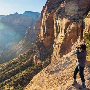 A person stands on a rocky cliff with a camera, capturing a view of a vast canyon landscape bathed in sunlight. The scene features rugged cliffs and a lush, sprawling valley.