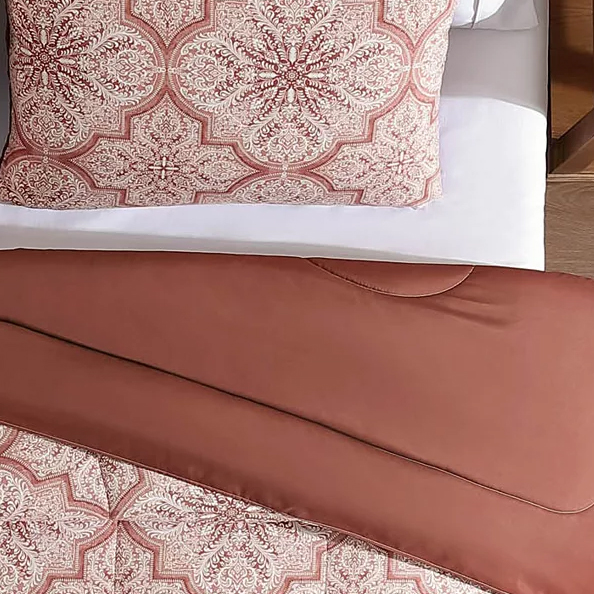 A decorative pillow and matching comforter feature an intricate, symmetrical floral pattern with a rust and beige color scheme. The bedding is arranged on a white sheeted bed.