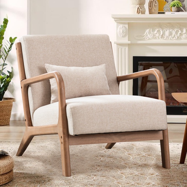 A beige upholstered armchair with wooden frame and armrests, accompanied by a small matching pillow, set in a cozy living room with a decorative fireplace and plants.