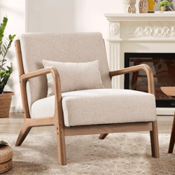 A beige upholstered armchair with wooden frame and armrests, accompanied by a small matching pillow, set in a cozy living room with a decorative fireplace and plants.