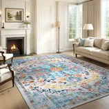 A living room with a large, colorful area rug featuring a vibrant floral and abstract pattern. The space includes a fireplace, cream-colored sofa, light armchair, and standing lamps set against a backdrop of large windows with beige curtains.