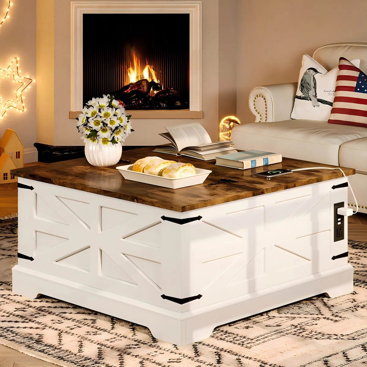 A white farmhouse-style lift-top coffee table with a wooden top, featuring decorative accents and built-in outlets. It holds a vase of flowers, a book, and a tray with bread, set in a cozy room with a fireplace.