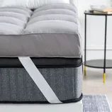 A 4-inch down-alternative mattress topper with quilted gray fabric and corner straps is layered on a mattress, providing added cushioning and support. A round side table with a book and mug is partially visible beside the bed.