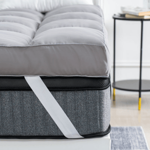 A 4-inch down-alternative mattress topper with quilted gray fabric and corner straps is layered on a mattress, providing added cushioning and support. A round side table with a book and mug is partially visible beside the bed.