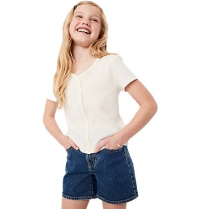 A young girl is wearing a short-sleeved, ribbed, white button-up top with high-waisted denim shorts, accessorized with a colorful bracelet.