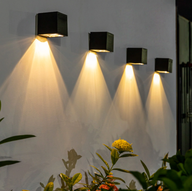 Four wall-mounted outdoor lights projecting downward beams on a white wall, with plants at the bottom.