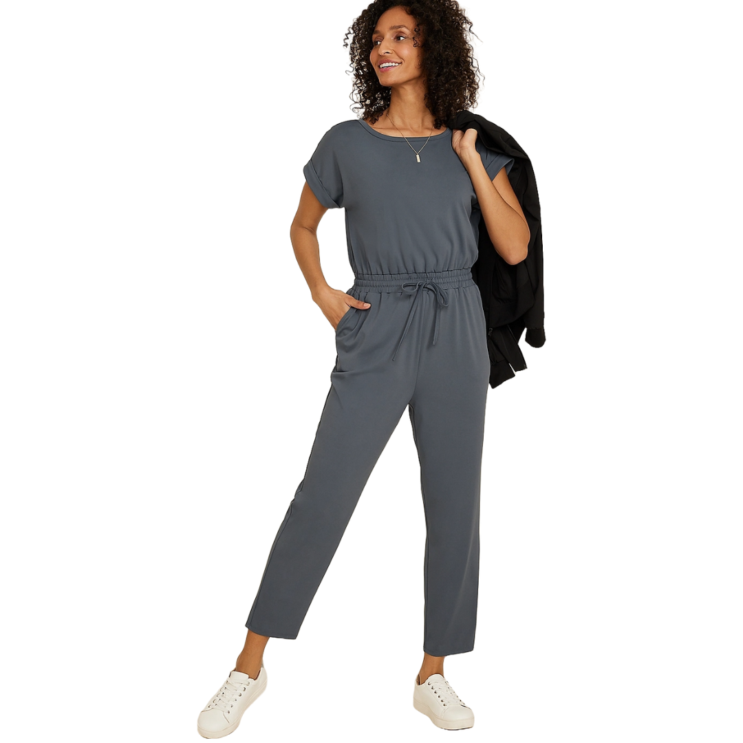 A woman wears a gray, short-sleeve Maurices Traveler Jumpsuit with a drawstring waist and ankle-length pants. She holds a black jacket and wears white sneakers, accessorized with a simple necklace.