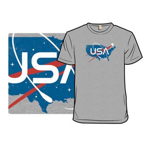 Grey T-shirt featuring the text \“USA\“ with a stylized design resembling a space agency logo. The graphic includes a map of the United States, a red swoosh, and white orbital rings against a blue backdrop with stars.