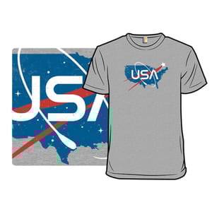 Grey T-shirt featuring the text \“USA\“ with a stylized design resembling a space agency logo. The graphic includes a map of the United States, a red swoosh, and white orbital rings against a blue backdrop with stars.