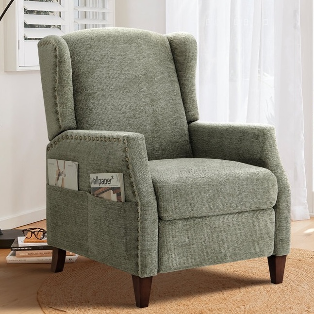 A green chenille push-back recliner featuring a high back with winged sides, nailhead trim, and wooden legs. The chair has side pockets for magazines and stands on a round woven rug.