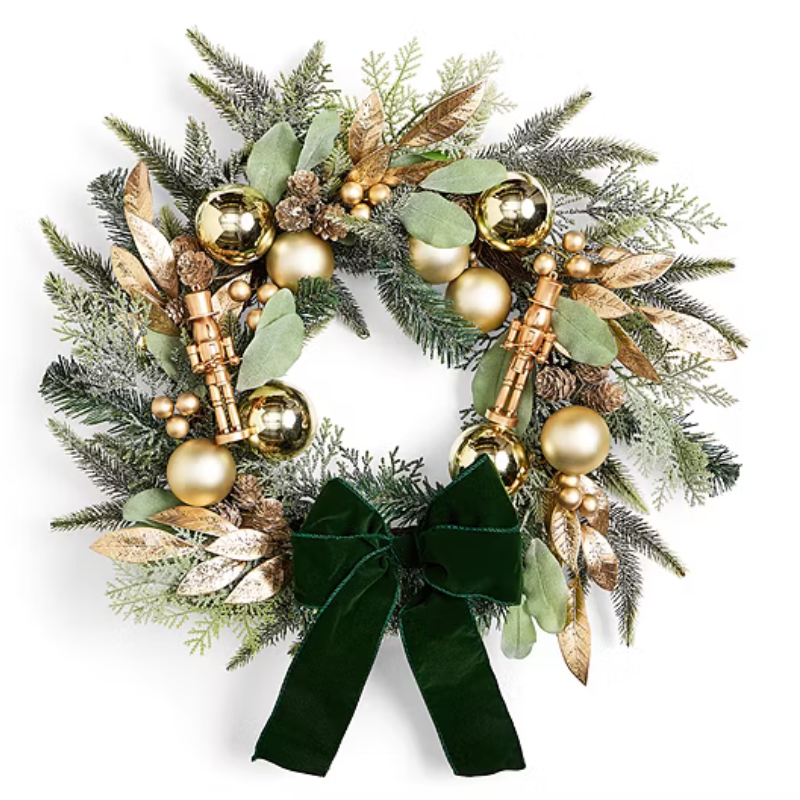 A decorative wreath featuring green and gold elements, including glossy spheres, leafy accents, small nutcracker figures, and a velvet green ribbon.