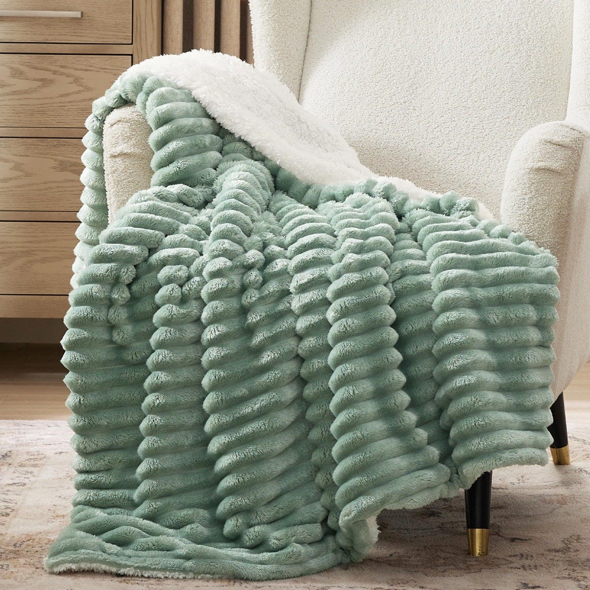 Plush mint green textured throw blanket with a white fleece underside, draped over a white armchair with black legs featuring gold accents, next to a wooden dresser.