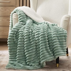 Plush mint green textured throw blanket with a white fleece underside, draped over a white armchair with black legs featuring gold accents, next to a wooden dresser.