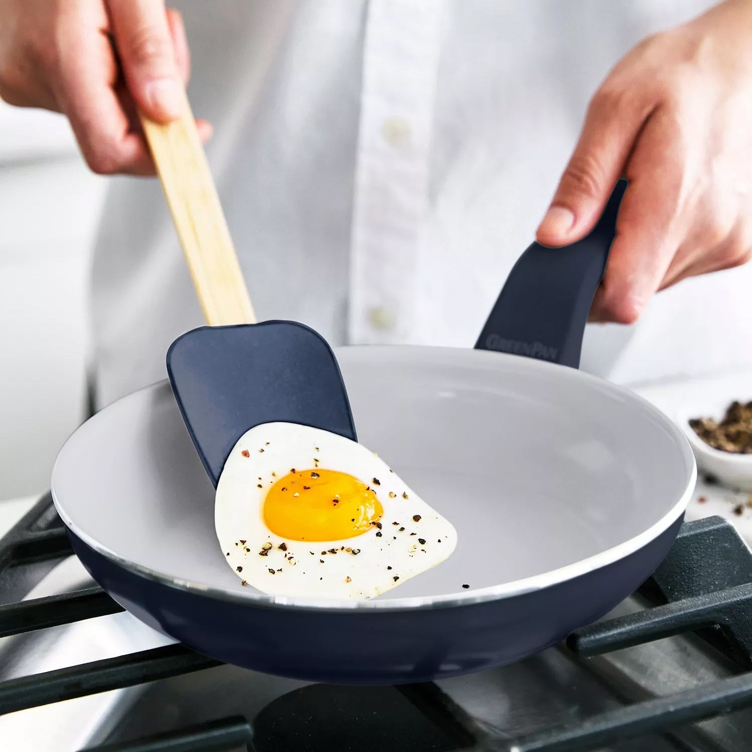 A person is using a frying pan with a non-stick surface to cook a sunny-side-up egg, using a blue spatula.