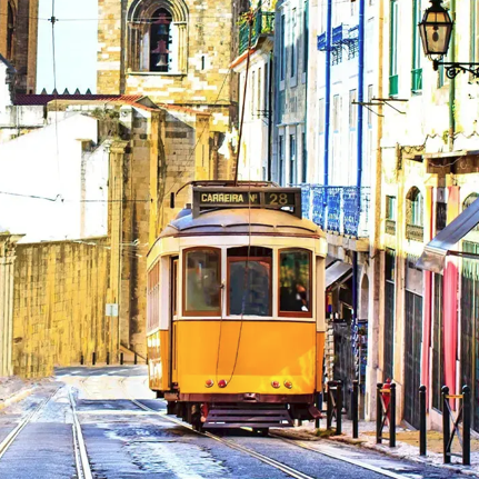 A yellow tram navigates a narrow, cobblestone street lined with colorful buildings, set against the backdrop of ancient architecture and a clear blue sky.