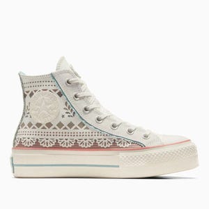 Converse Day of the Dead shoes feature a high-top design with intricate lace-like patterns, floral motifs, and a blend of soft pastel colors including white, pink, and blue, all set on a platform sole.