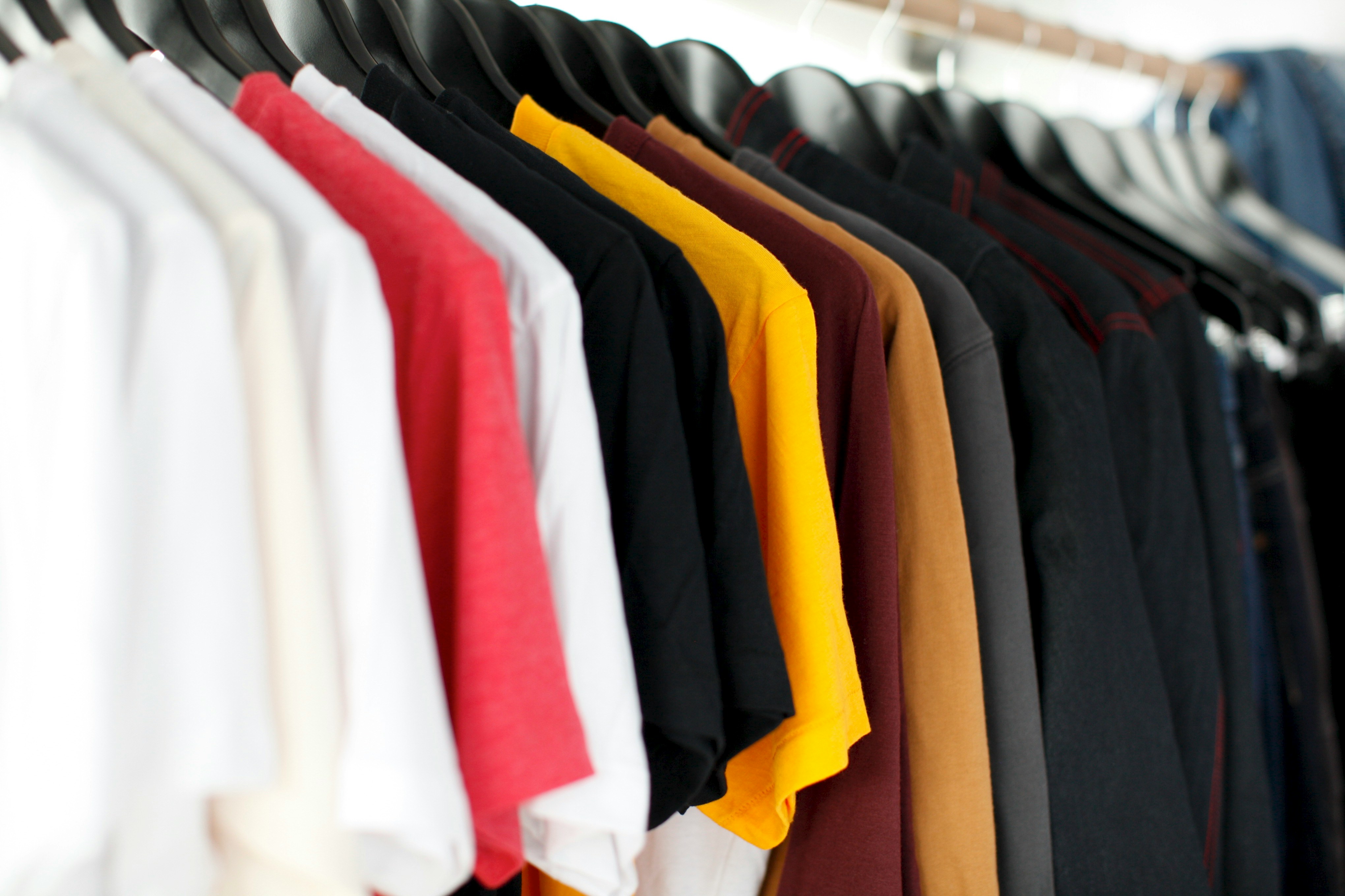A selection of T-shirts in various solid colors hung on a rack, alongside darker jackets and denim jeans.