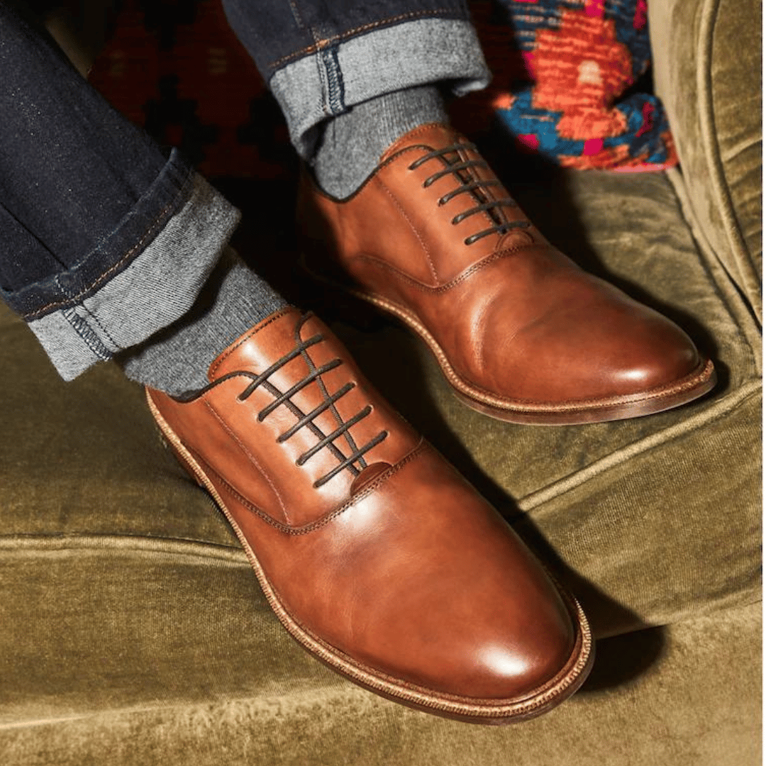 Brown leather dress shoes with laces, paired with gray socks and rolled-up dark denim jeans.