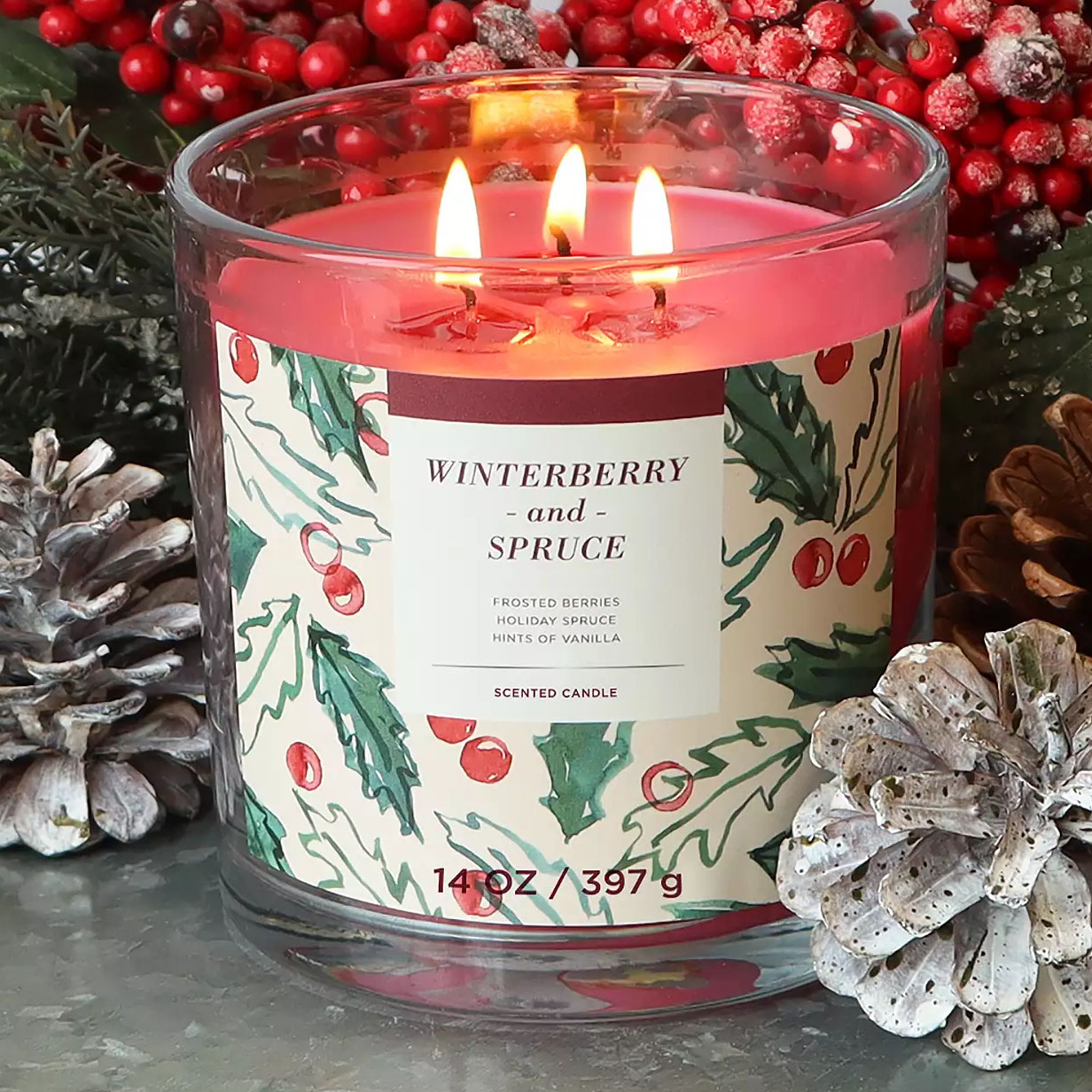 A 14 oz scented candle labeled \“Winterberry and Spruce\“ with notes of frosted berries, holiday spruce, and hints of vanilla is surrounded by pinecones and red berries.