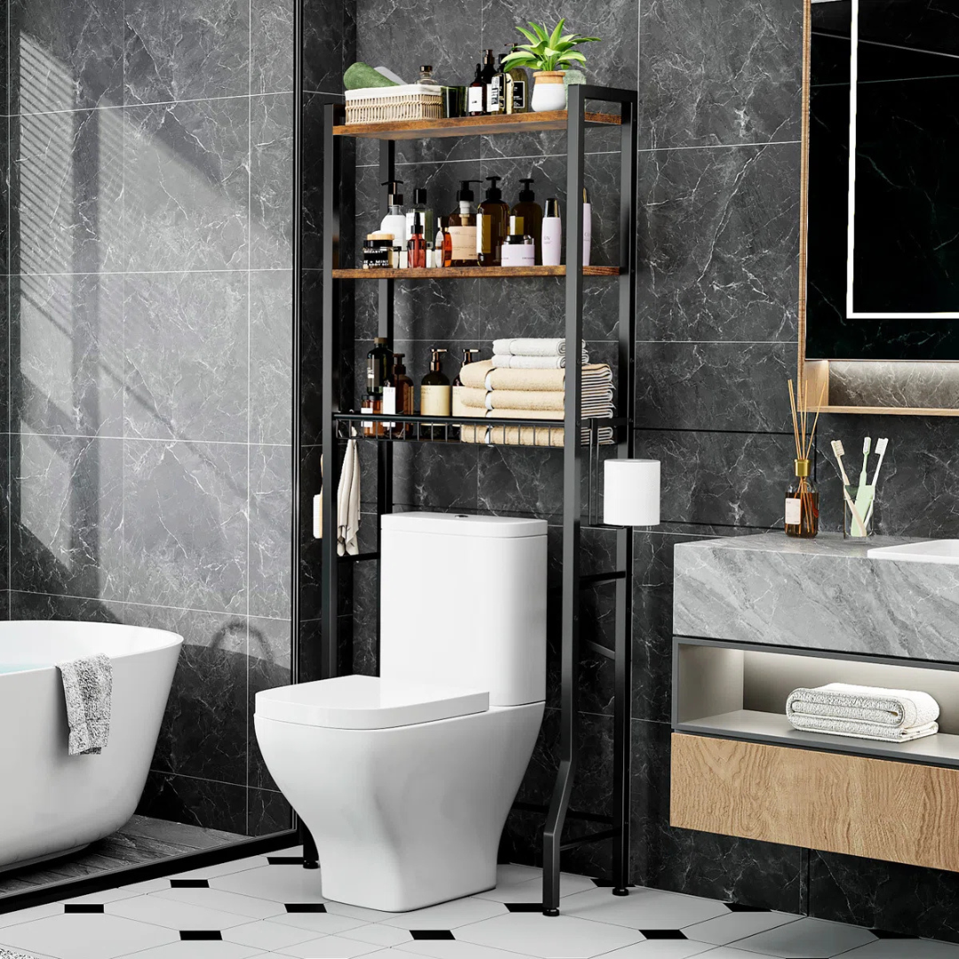 A bathroom features a toilet with a black metal over-the-toilet storage rack holding toiletries, towels, and a plant. Nearby are a bathtub, a marble countertop with a wooden drawer, a mirror, and a toothbrush holder.