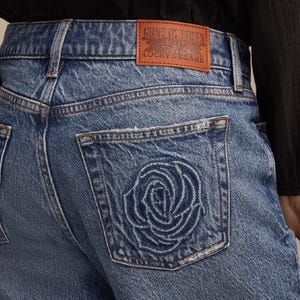 Denim jeans featuring a rose design on the back pocket and a leather patch labeled \“Guns N' Roses Lucky Brand.\“