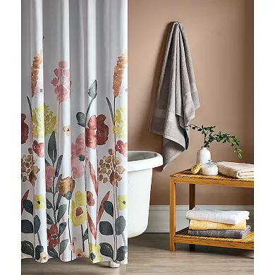 A grey bath towel is casually hung over a towel rack in a bathroom setting, paired with a floral shower curtain and wooden furniture displaying neatly folded white towels.