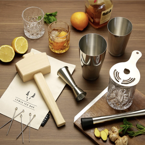Cocktail making set with a Lewis ice bag, wooden mallet, jigger, shaker tins, strainer, skewers, and garnish items like lemons, ginger, and mint alongside a cutting board and a bottle of spirits.