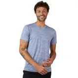 A man wearing a light blue short-sleeve T-shirt with a subtle heather pattern stands against a gray background. He has several tattoos on his arms and is smiling.