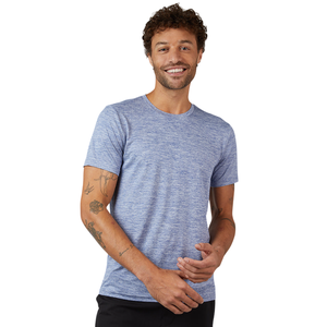 A man wearing a light blue short-sleeve T-shirt with a subtle heather pattern stands against a gray background. He has several tattoos on his arms and is smiling.