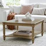 A wooden square coffee table with turned legs features a shelf below. It is styled with books, a white abstract sculpture, and a mug. The background includes a beige couch with checkered and striped pillows and a brown throw blanket.